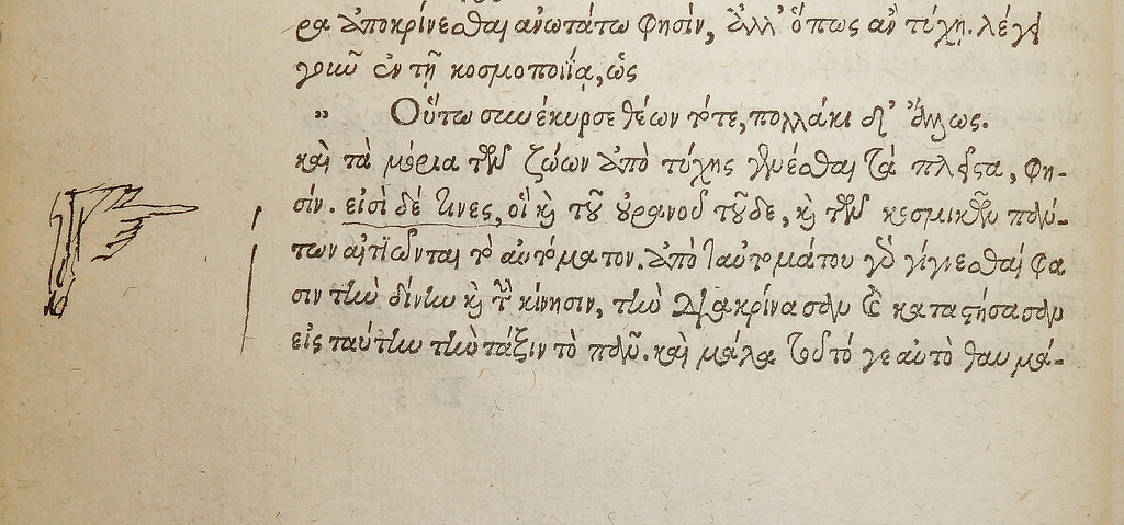 Una mano disegnata a inchiostro nel margine di un libro in greco, con l'indice puntato su una riga.